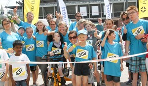 Eine fröhliche Gruppe von Erwachsenen und Kindern mit und ohne Behinderungen  in hellblauen T-Shirts hinter der Startlinie