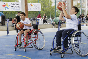 zwei Jungen in Sollstühlen spielen Basketball im Hintergrund ein Banner mit der Aufschrift „SportWoche für Alle“