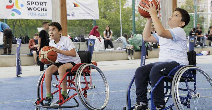 zwei Jungen in Sollstühlen spielen Basketball im Hintergrund ein Banner mit der Aufschrift „SportWoche für Alle“