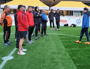 ein Mann (Taime Kuttig) steht auf einem Fußballfeld vor einer Gruppe von Männern in Sportkleidung
