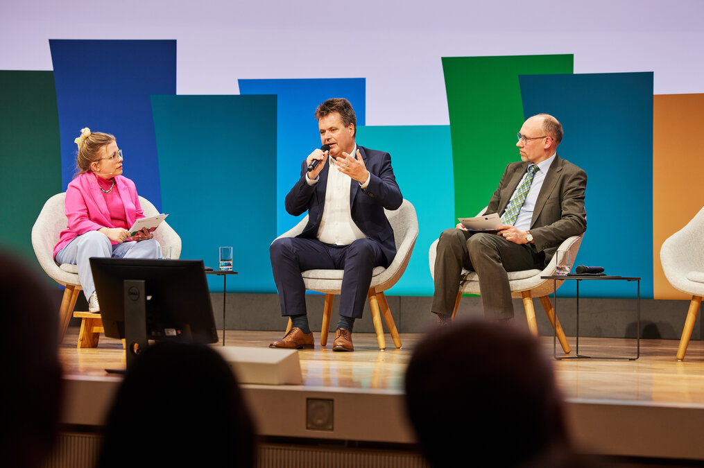 drei Personen sitzen auf dem Podium und diskutieren, Jürgen Dusel, die person in der Mitte spricht gerade und gestikuliert dabei drei Personen sitzen auf dem Podium und diskutieren, Jürgen Dusel, die person in der Mitte spricht gerade und gestikuliert dabei