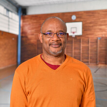 ein Mann im orangefarbenen Pullover, im Hintergrund das Bild einer Sporthalle in ähnlichen Farbtönen