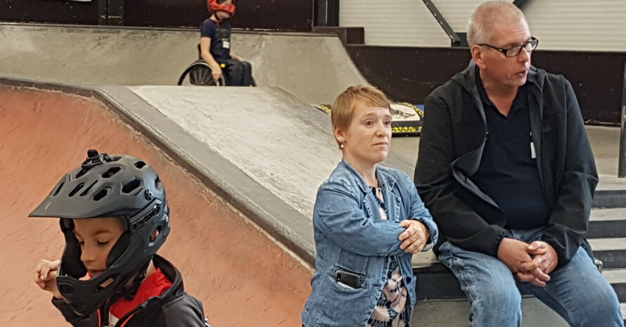 eine kleinwüchsige Frau und ein Mann inmitten eines Skateparks hinter einem Kind mit coolem Helm und Rollstuhl im Vordergrund