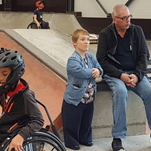 eine kleinwüchsige Frau und ein Mann inmitten eines Skateparks hinter einem Kind mit coolem Helm und Rollstuhl im Vordergrund