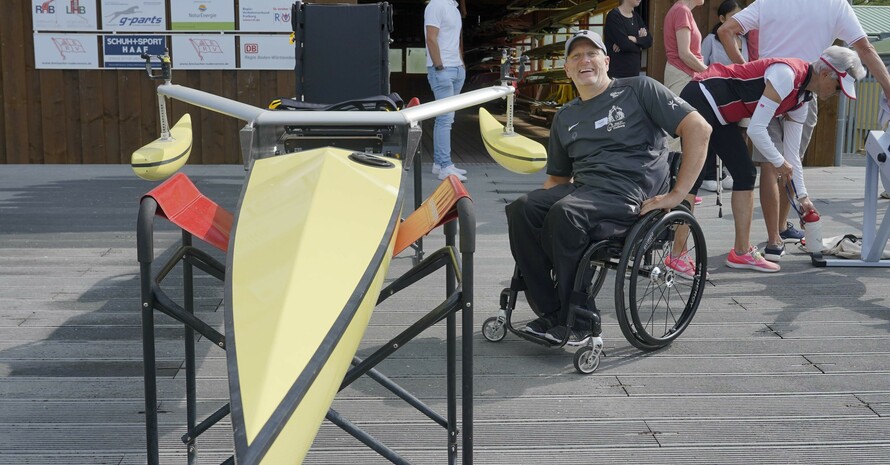Eine lachende Person, die im Rollstuhl sitzt,  befindet sich rechts neben einem Para Ruderboot. Das umgedreht Para Ruderboot liegt auf zwei Böcken. Im Hintergrund stehen weitere Personen