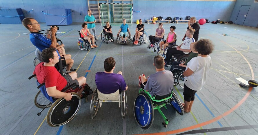 eine Gruppe von Rollstuhlfahrer*innen bildet in einer Turnhalle einen Kreis, drei Personen stehen außerhalb, einer davon fotografiert, die anderen erklären 