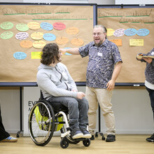 Vier Personen, einer von ihnen im Rollstuhl, diskutieren vor einer Pinnwand mit farbigen Karten