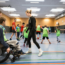 eine Gruppe von Kindern und Jugendlichen spielt in einer Sporthalle Fußball im Vordergrund zwei Personen und ein Rollstuhlfahrern