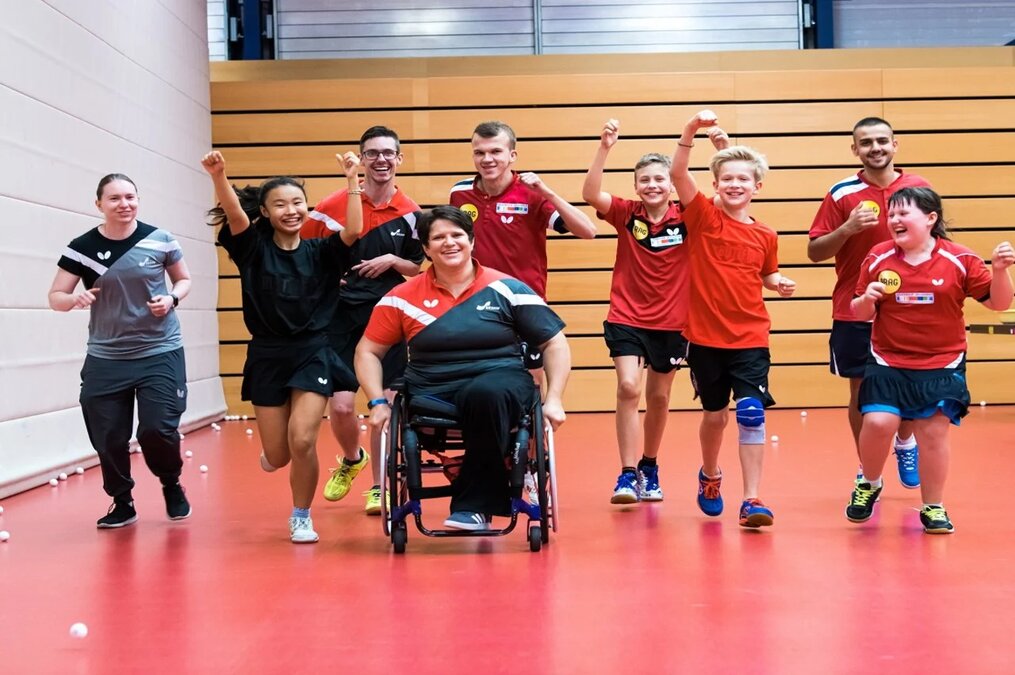 Eine Gruppe von Sportler*innen unterschiedlichen Alters läuft mit viel Spaß in einer Sporthalle, im Vordergrund eine Rollstuhlfahrerin
