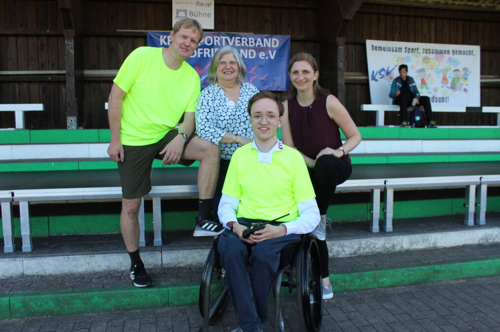 Vier Personen vor einem Plakat des Kreissportverbands Nordfriesland. Die beiden Männer tragen leuchtend gelbe T-Shirts, die sie als Mitarbeitende kennzeichnen, einer davon, Event-Inklusionsmanager Tyll Reinisch sitzt im Rollstuhl. Dahinter zwei Frauen. Vier Personen vor einem Plakat des Kreissportverbands Nordfriesland. Die beiden Männer tragen leuchtend gelbe T-Shirts, die sie als Mitarbeitende kennzeichnen, einer von beiden, Event-Inklusionsmanager Tyll Reinisch sitzt im Rollstuhl, stehend dahinter Tade Corinth und die für das Projekt zuständigen Mitarbeiterinnen